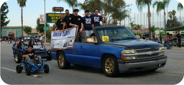 tudents ride in and stand around a blue pickup truck during a Buckboard Day parade. A student in a football jersey holds a Rowland Raiders banner, while another student sits in a small blue go-kart nearby. Palm trees, storefronts, and spectators line the street.