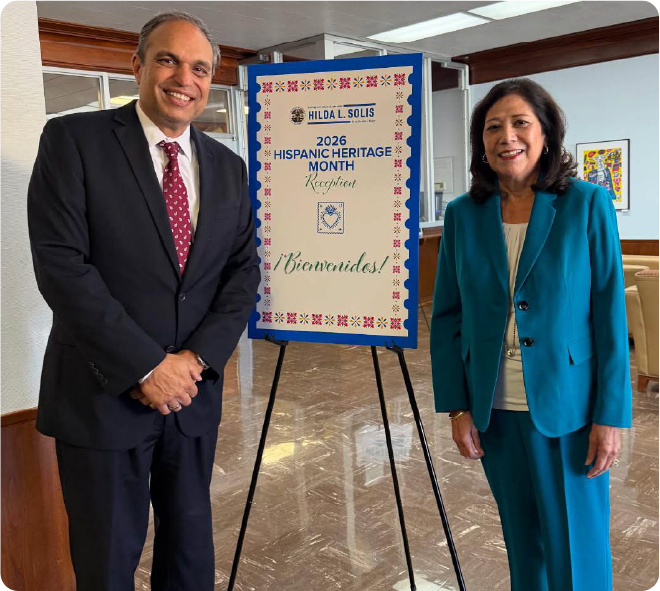 Two adults stand beside a decorated sign for the 2026 Hispanic Heritage Month Reception, which features colorful floral borders and the words “¡Bienvenidos!” in the center.