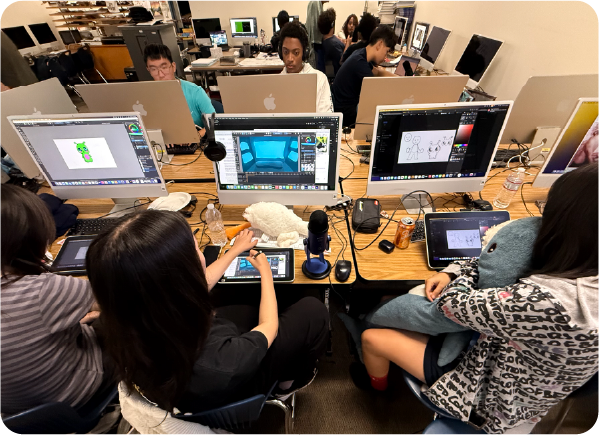 Students work at computers in a classroom equipped with large monitors, drawing tablets, and creative software. Several screens display digital illustrations and graphic design projects as students collaborate at their desks.
