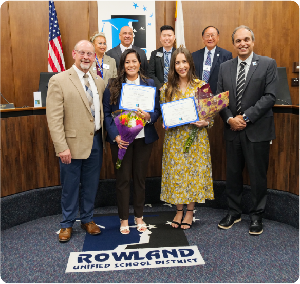 Two educators hold certificates and bouquets of flowers during a recognition ceremony with school district administrators.