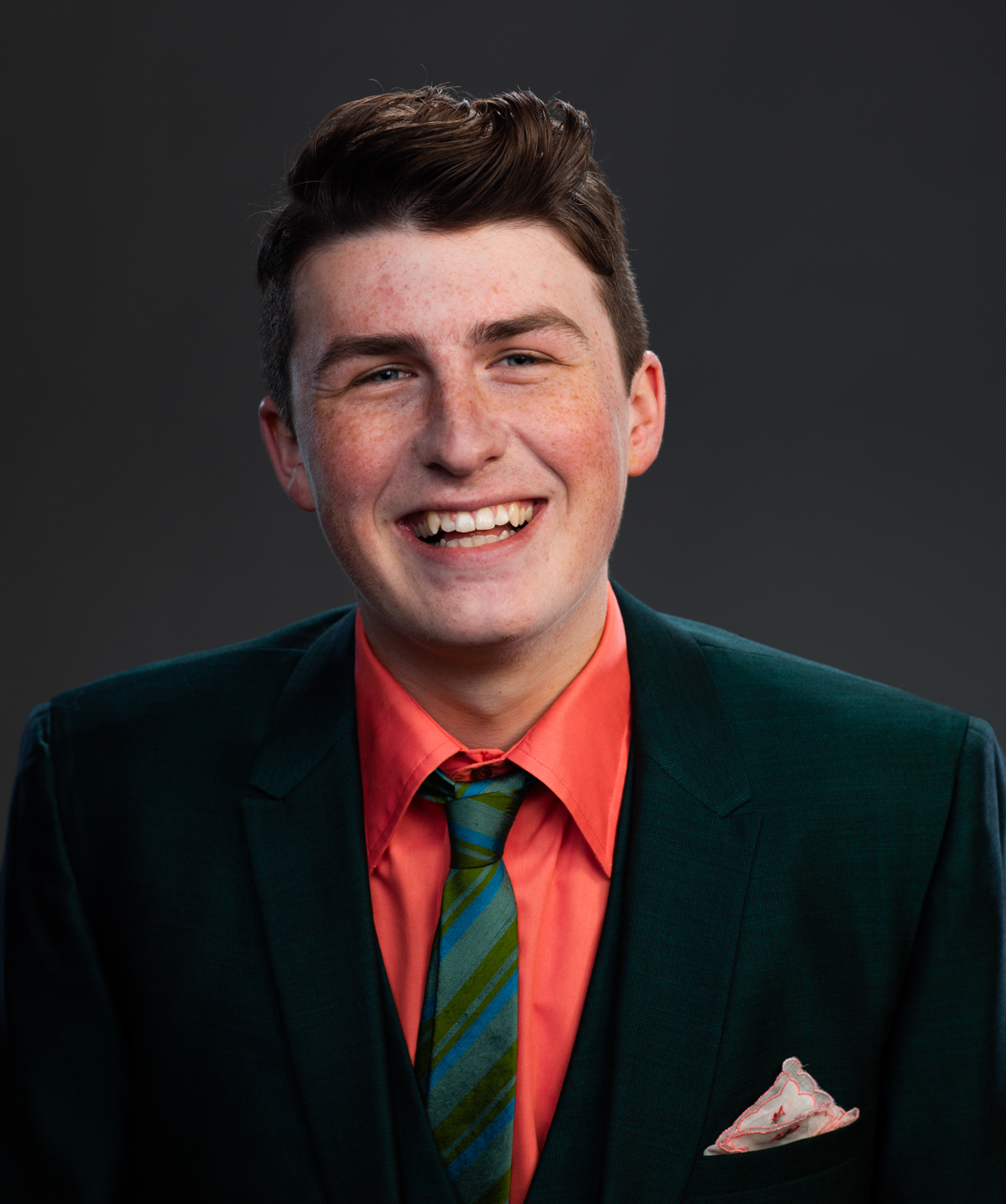 a white man with brown hair smiling into the camera. He is wearing a green suit jacket an orange collared shirt and green tie. 