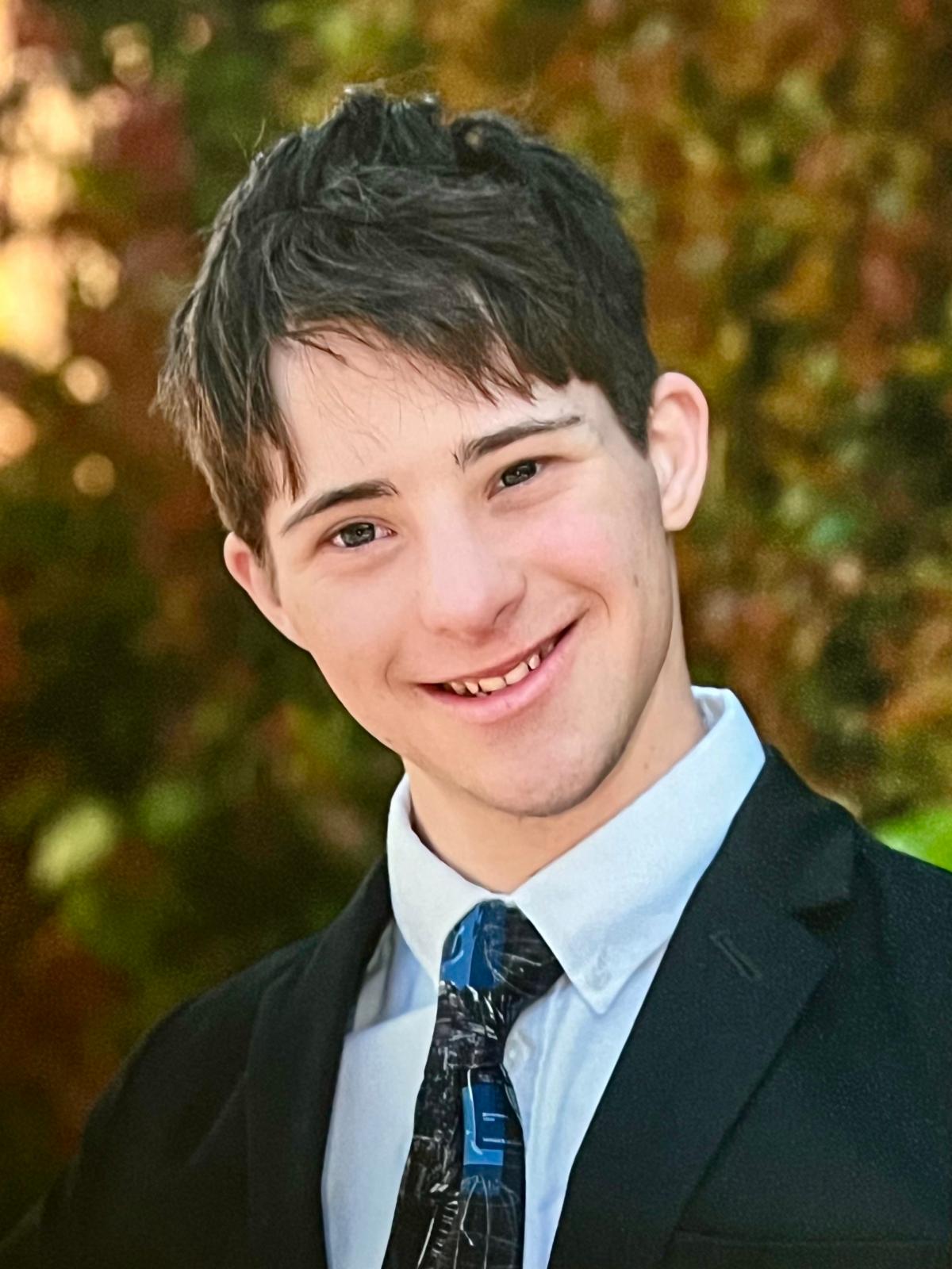 a white man with brown hair smiling into the camera. He is wearing a black suit jacket a blue collared shirt and black and blue tie. 