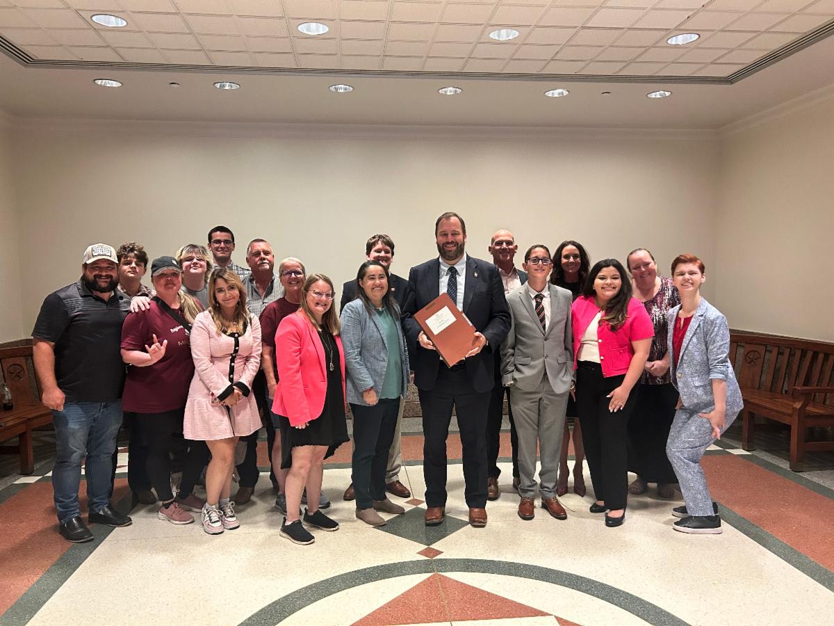 A group of approximately 20 people of diverse ages and backgrounds stand together indoors smiling at the camera. The central figure a man in a dark suit holds a large folder. The group is standing in a well-lit room with high ceilings and decorative floori