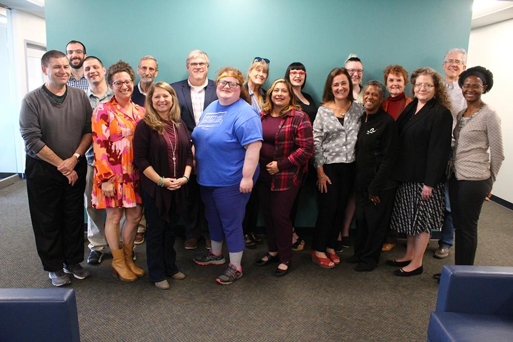 A group of 18 adults stands together in an office smiling at the camera in front of a teal colored wall. The group includes people of different ages and genders and races dressed in a mix of casual and business casual clothing.