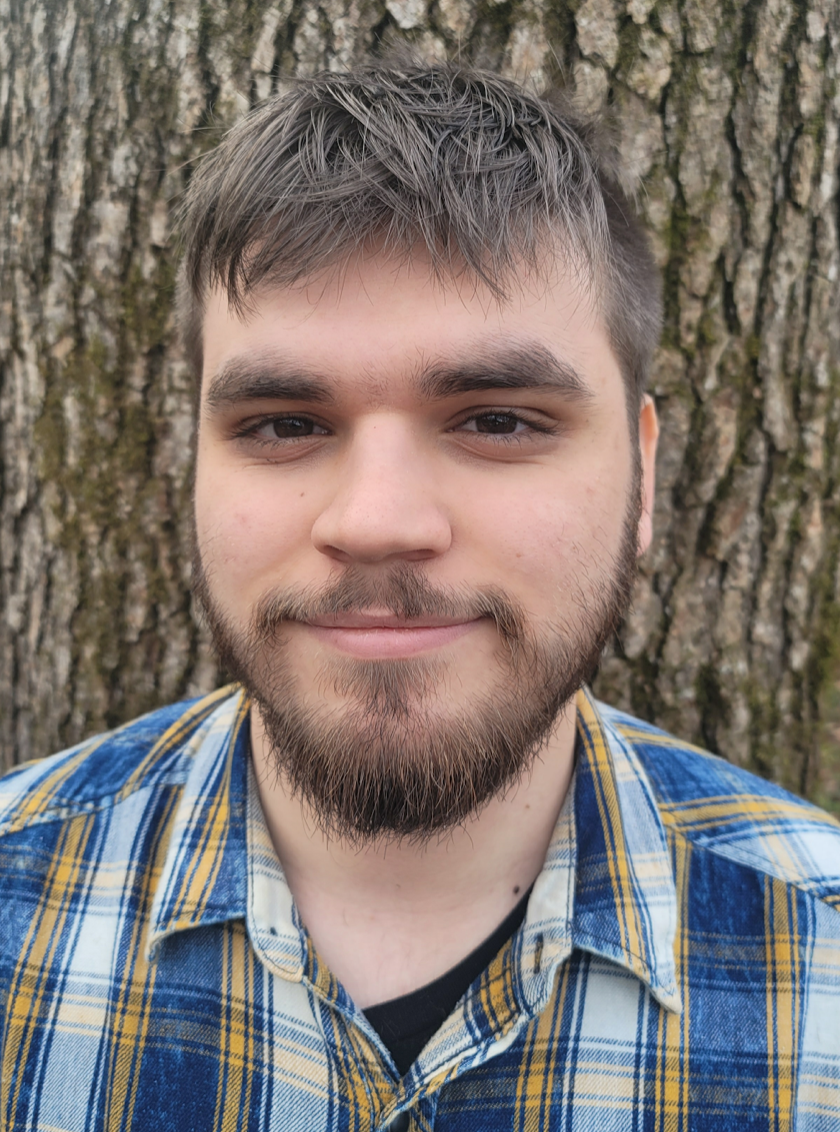 Photo of a light skinned man with a beard in front of a tree