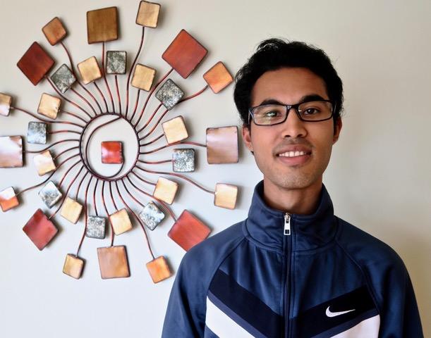 a man with dark skin and brown hair smiling into the camera. He is wearing black framed glasses and a blue Nike sweatshirt. To his left is a wall ornament of various earth tones.