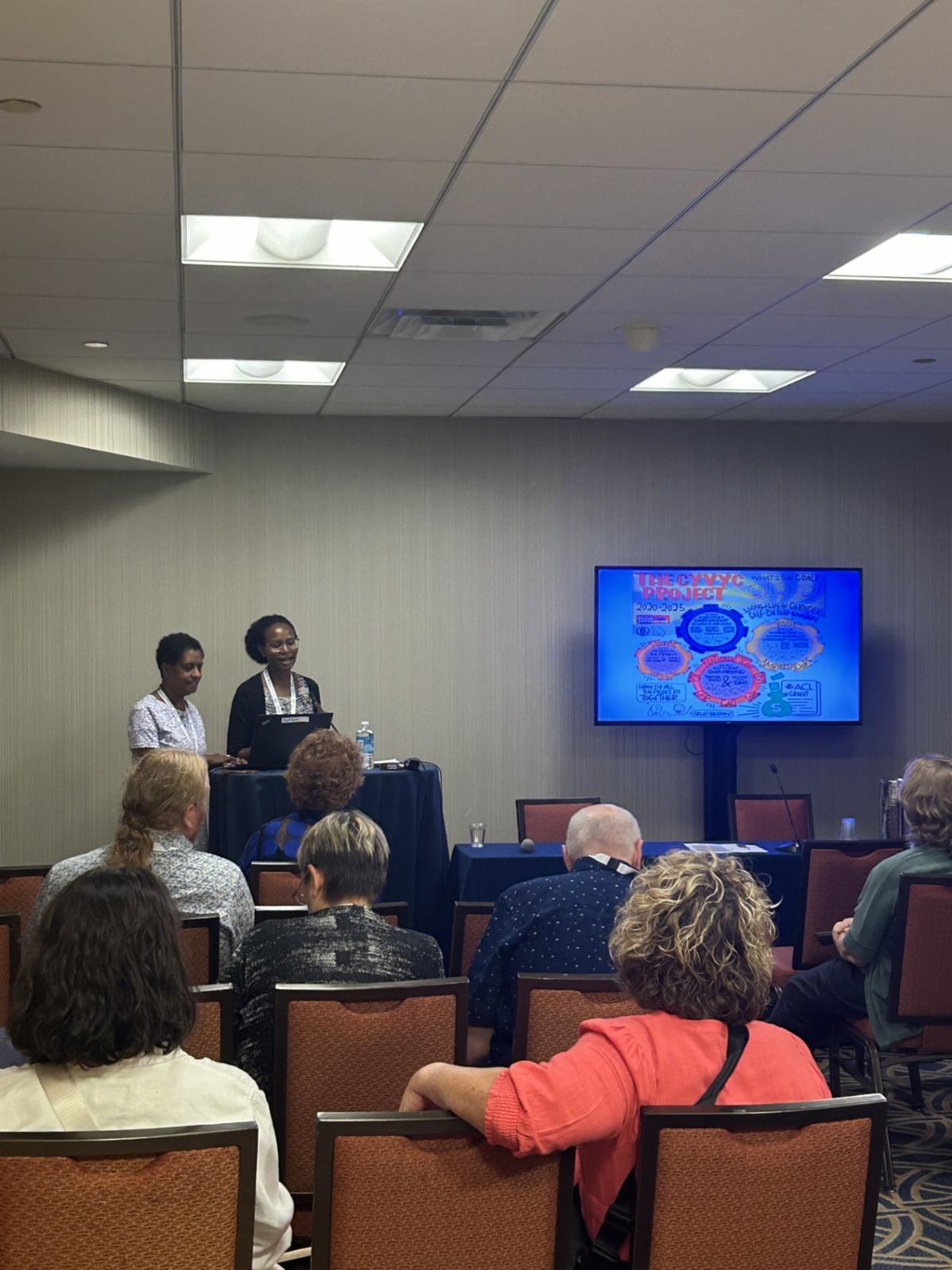Two people present at the front of a room next to a tv screen