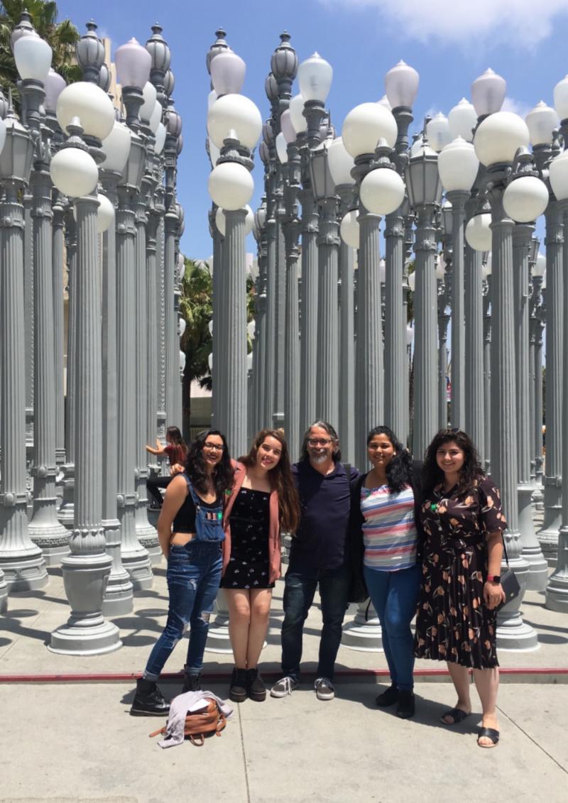 Four female students. Professor at the center  with students posing for the camera. Light posts behind them. 