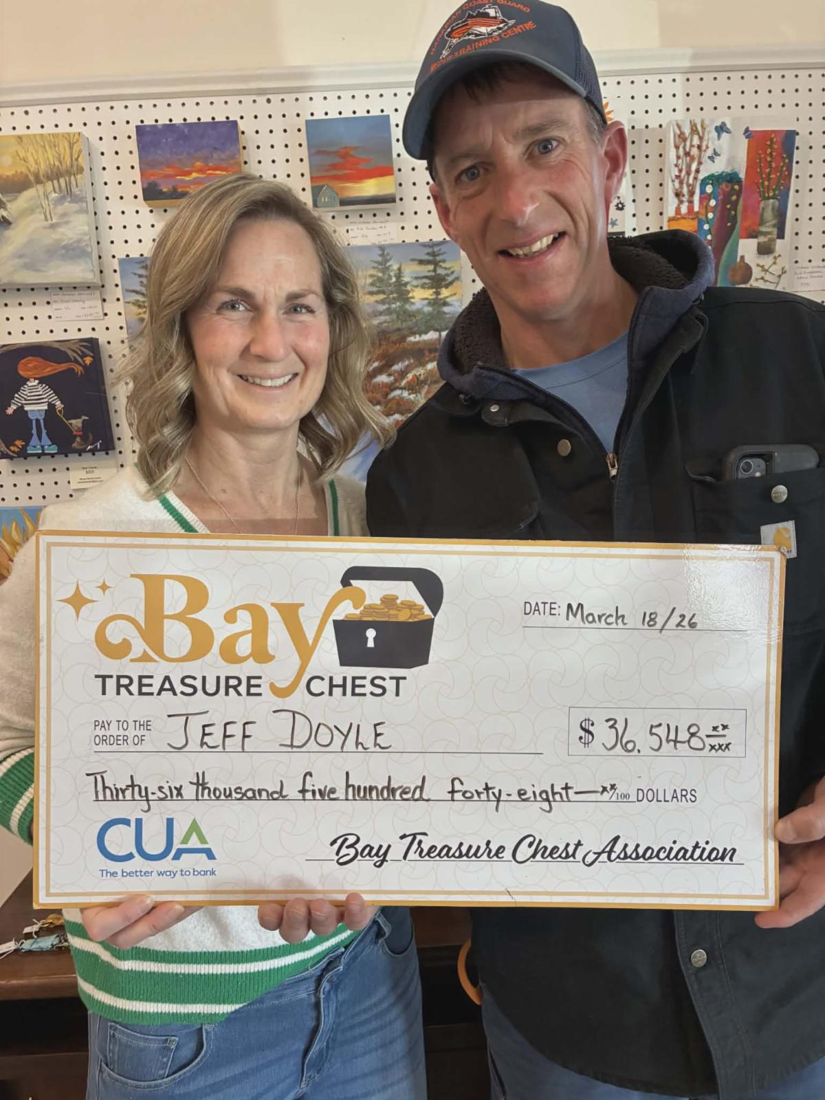 woman wearing a white sweater with a man wearing a black jacket and ballhat holding an oversized cheque 