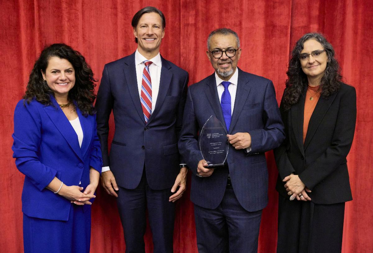 HHI and GHP faculty standing with award recipient who is holding a commemorative plaque, standing in front of a red curtain backdrop in business clothing