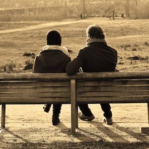 Sepia-tone photo of two adults sitting on a park bench as seen from behind looking out at a grassy park