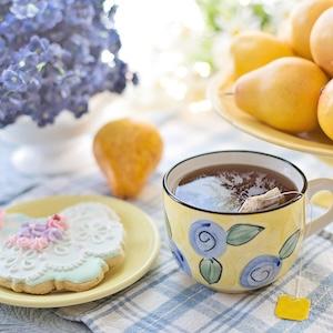 Stock photo of hot tea in a yellow mug with blue flowers next to a decorated sugar cookie on a table with fresh fruit and blue flowers