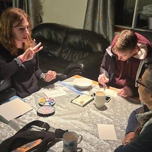 Photo of three people creating small watercolor paintings at a kitchen table