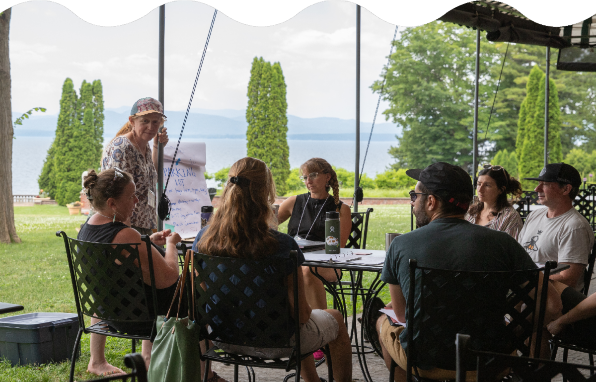 A group of people sit around a table outdoors, engaged in discussion. One person stands, presenting information on a flip chart.