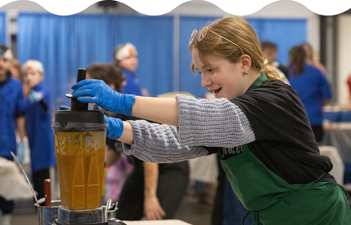 a young person smiles and holds the top of a blender while pureeing an orange liquid.
