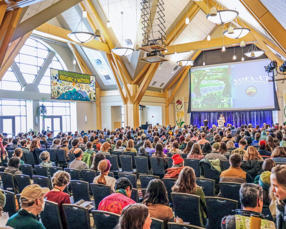   Audience attending a lecture at a NOFA-VT (Northeast Organic Farming Association of Vermont) conference, with a speaker visible in the distance under large wooden beams and informational banners.