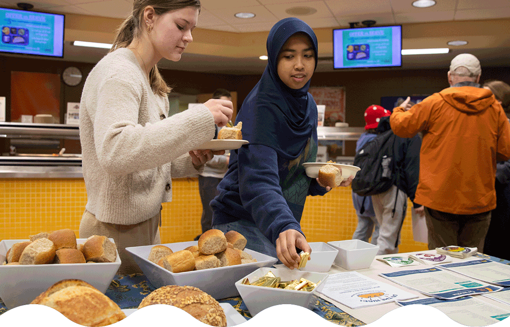  Generate by AI Assist  A group of people is gathered around a table in a cafeteria. Two individuals are serving themselves from trays of bread rolls and packets of butter. 
