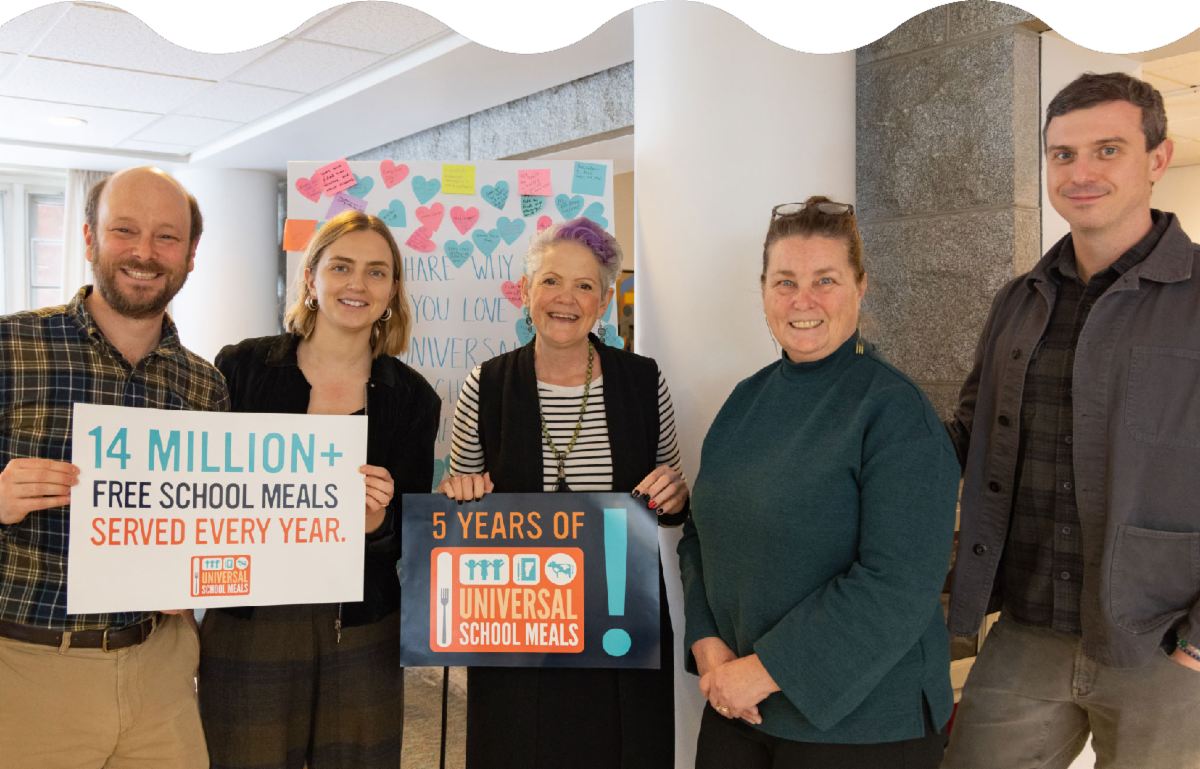 Five people stand together with posters. One posters reading '14 Million+ Free School Meals Served Every Year,' and '5 Years of Universal School Meals.'