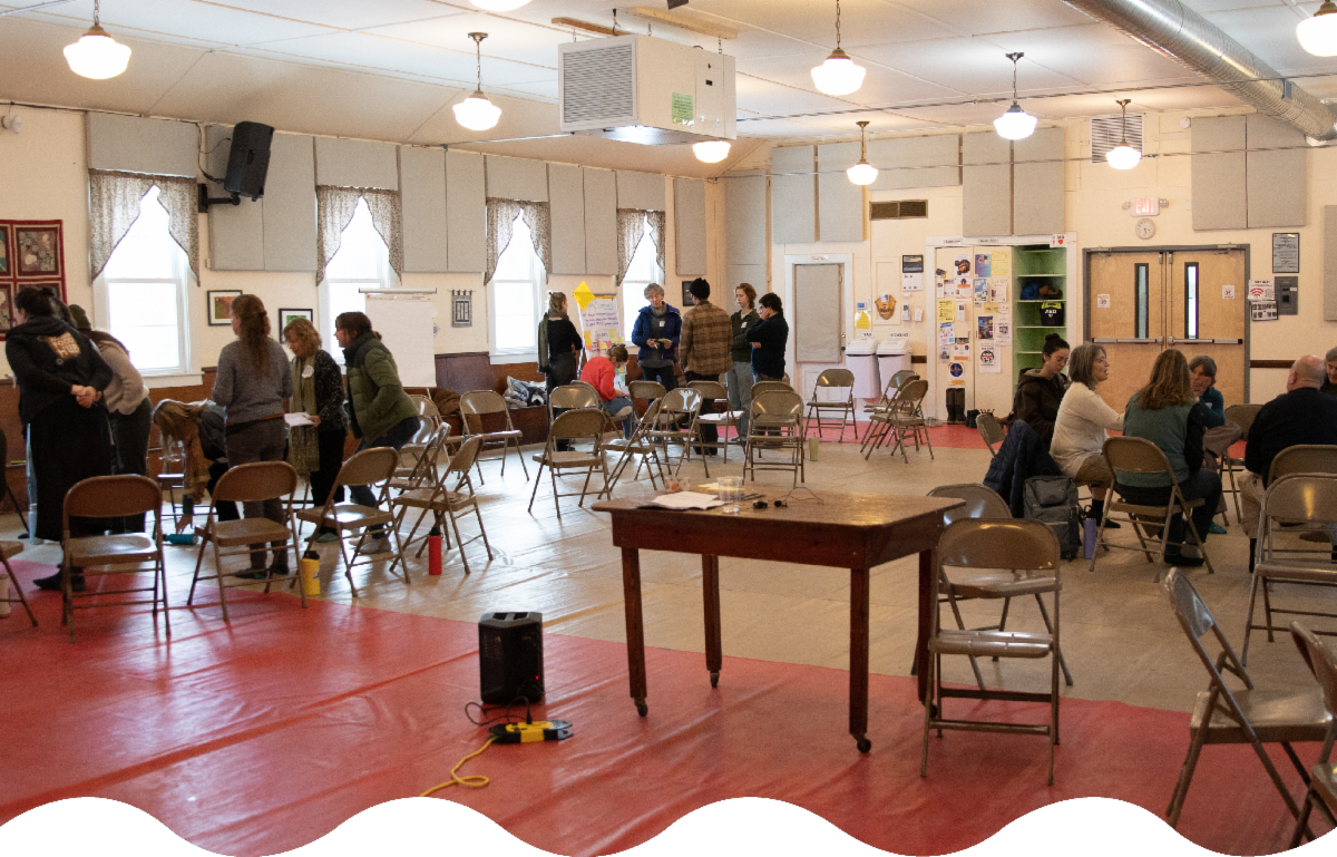 A community center room filled with people seated on folding chairs and engaged in conversation.