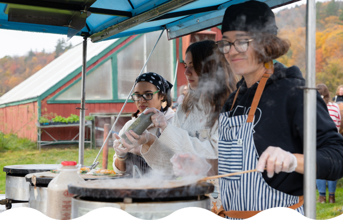 Three people cooking at an outdoor stall with a farm visible in the background.