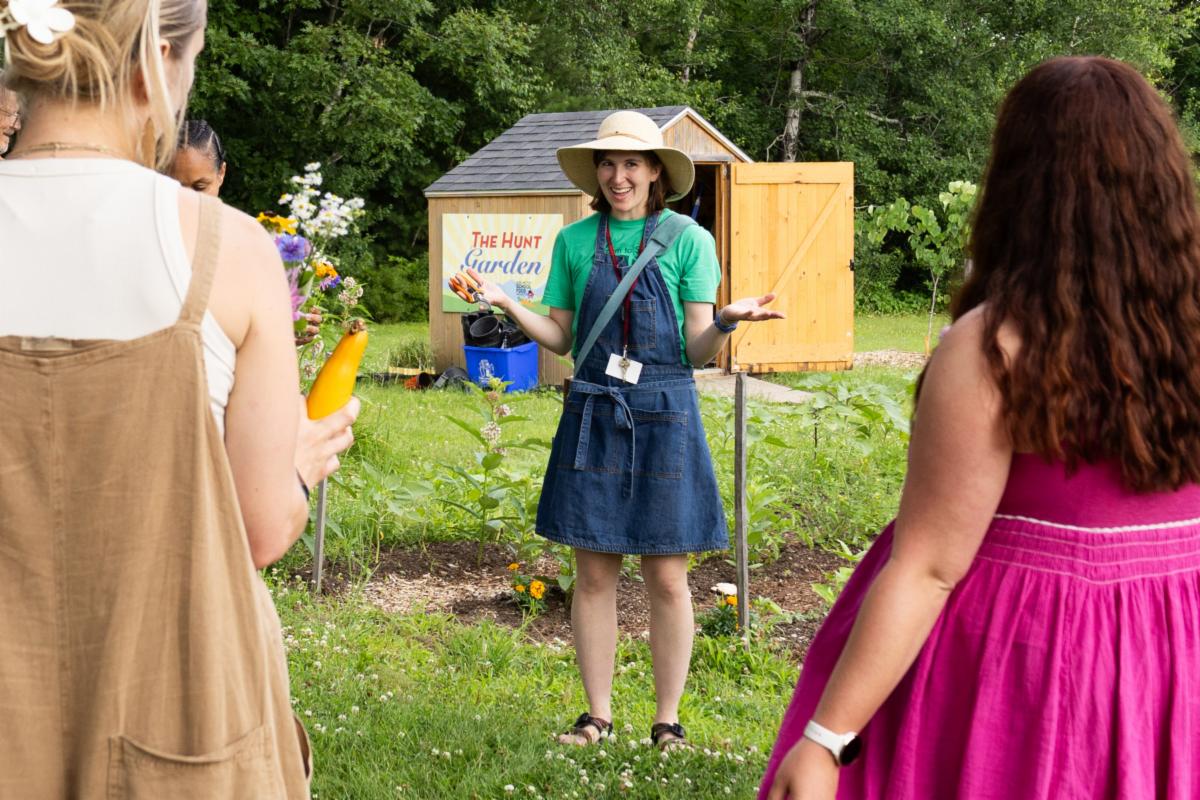 A person wearing a denim garden apron invites a group into an outdoor garden space.