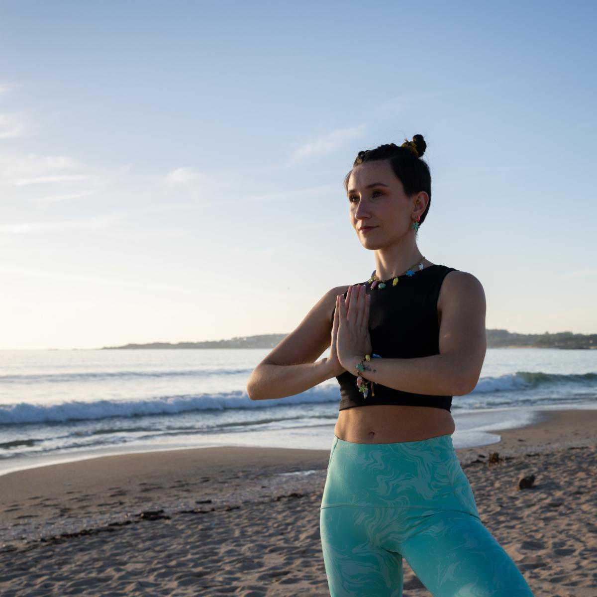 Young woman stands on a sandy beach with her hands in prayer position practicing yoga as the sun rises over the ocean creating a peaceful and serene atmosphere