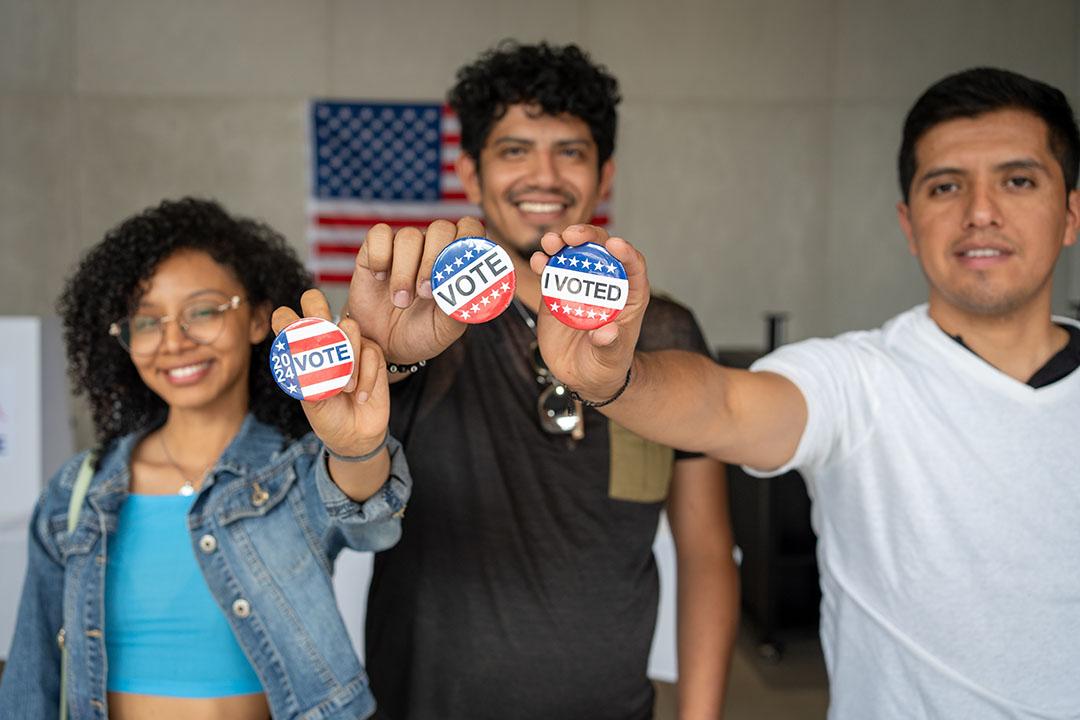 Three Hispanic smiling people holding I voted buttons and pins at an election polling station.
