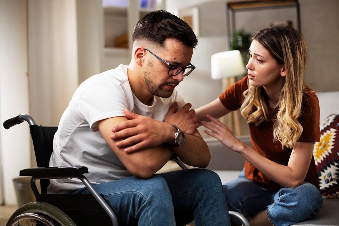 A young woman comforting a sad man sitting in a wheelchair with his head hanging down.