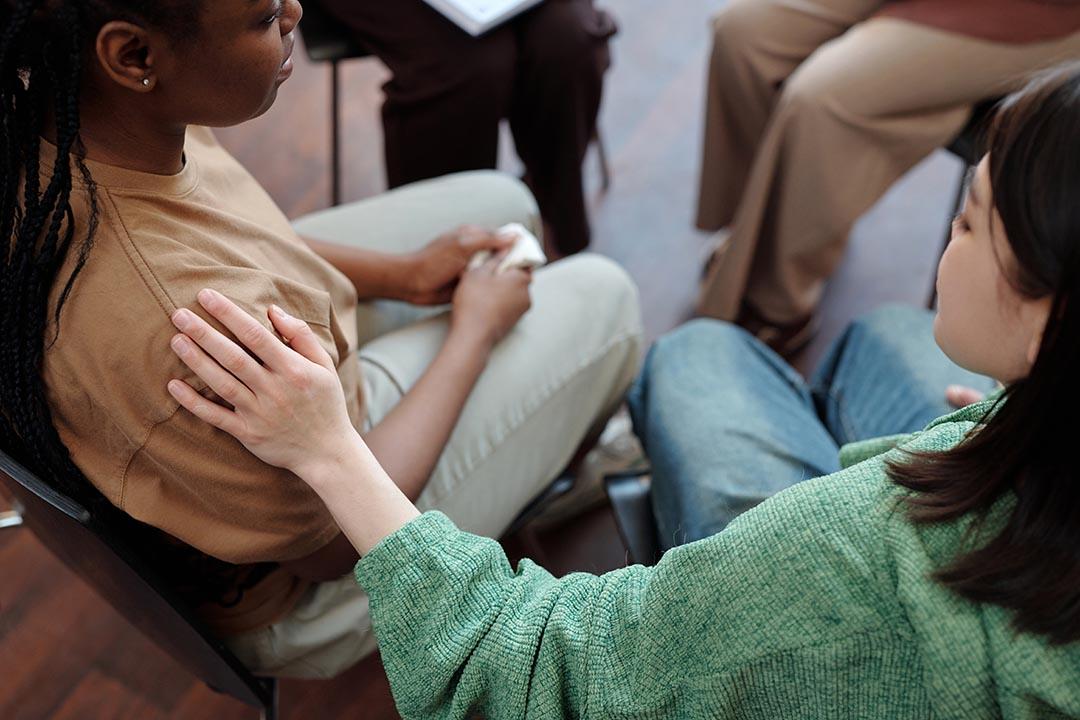 Asian woman seated comforting an African American woman sitting beside her with hand on her shoulder