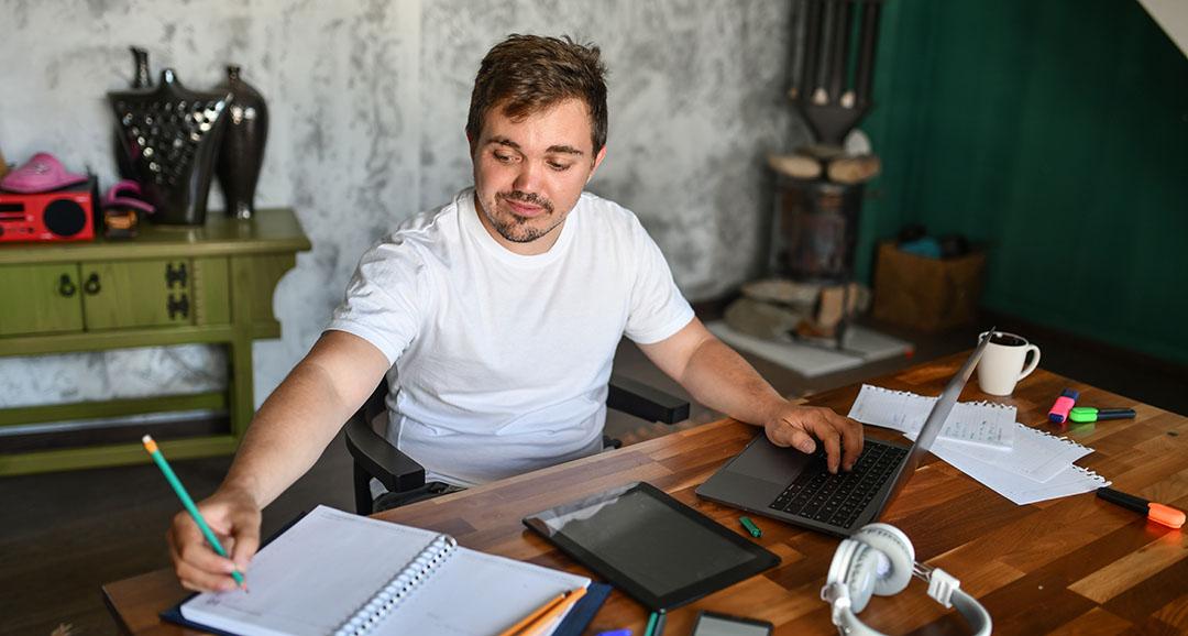 Man with dwarfism working remotely on a laptop computer at his home desk 