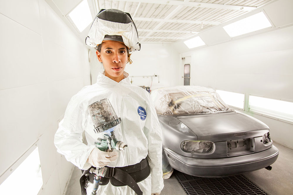 A student in the Auto Body and Paint Technician program wears a white jumpsuit and head covering while holding a paint gun. A silver car is in the background.
