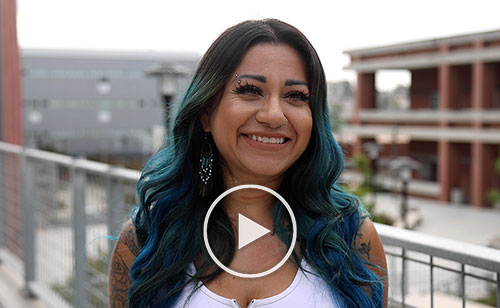 Nathaly Alvizures is smiling and wearing a white tank top and has black and teal hair. she is standing on a second story walkway with the campus behind her. 
