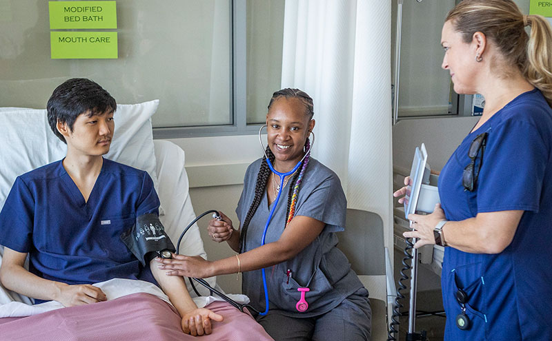 A medical student playing a patient is in a hospital bed having his blood pressure checked by another medical student while an instructor looks on.