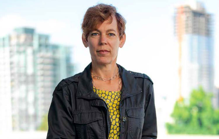 Anna Rogers stands on a balcony with the skyline of the city of San Diego behind her. She is wearing a yellow top and gray jacket.