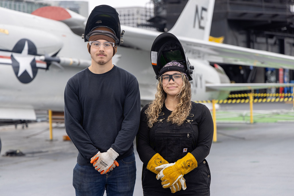 Two students in welding gloves and helmets on the USS Midway. There is a fighter plane in the background.