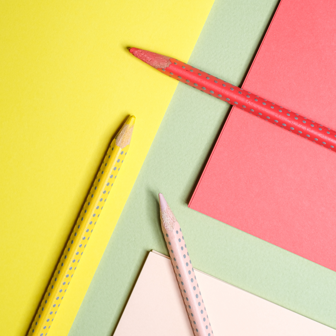 A bird's eye view photograph of 3 sheets of coloured paper (yellow, light pink and pink) on a green table with 3 pencils resting on top of the sheets. The pencils are yellow, pink, and red.