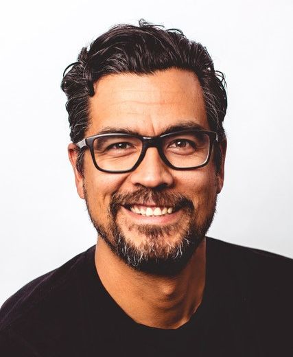 David Robertson has short, curly brown hair, black-rimmed glasses and is wearing a black T-shirt. He is smiling directly at the camera.