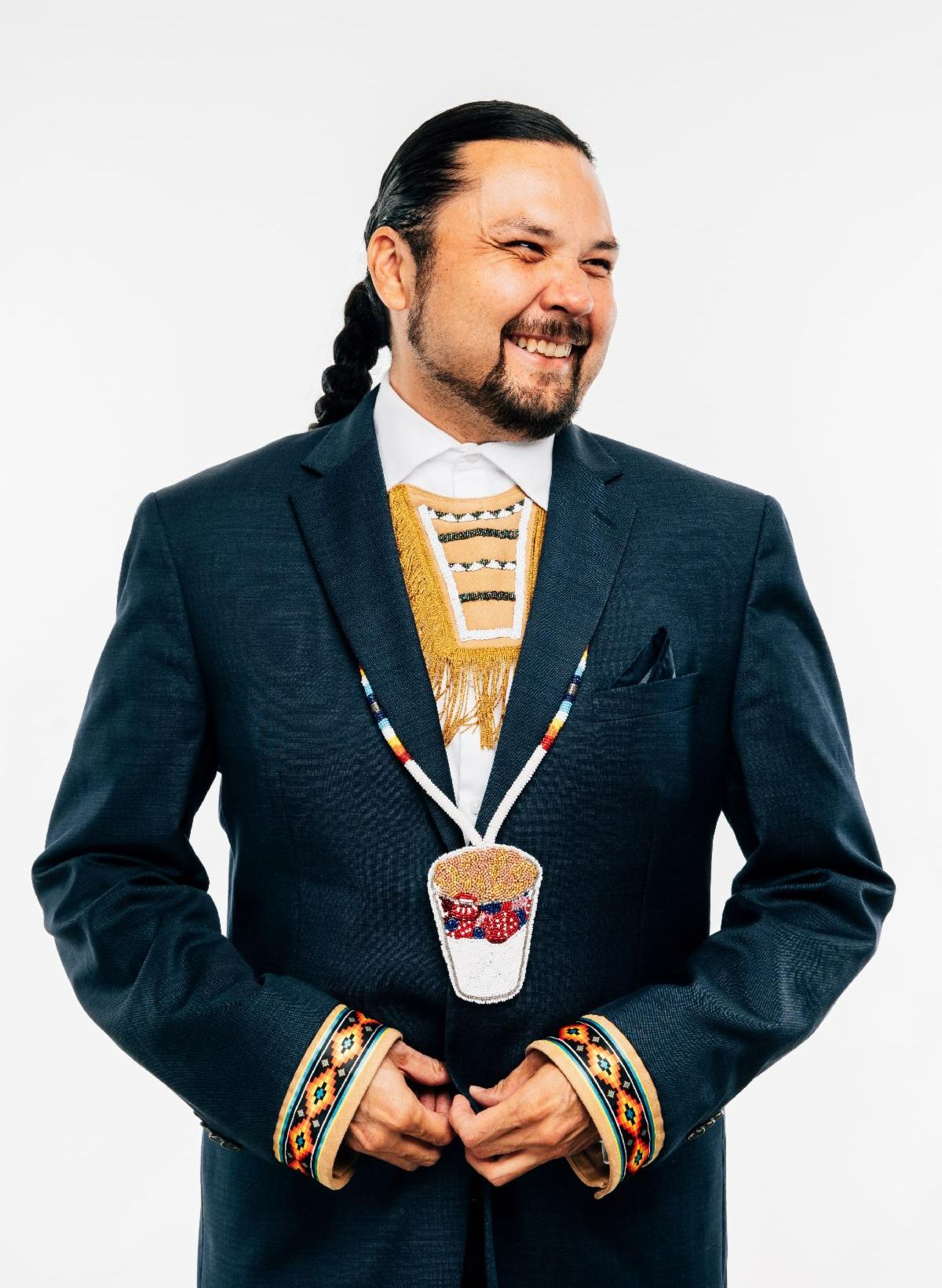 Shawn Cuthand has long black hair pulled back into a braid. He is wearing a navy blue suit with beaded cuffs and a beaded necklace. He smiles sideways at the camera.