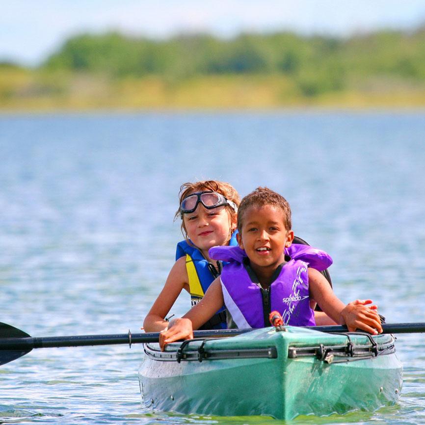 Two young girls wearing life jackets paddle a green kayak together. In the distance behind them, you can see the green of the grass and shrubs on the shore.