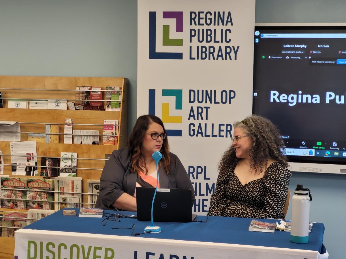 CBC's Sam Maciag does a live interview of Miss G and Me author Jennifer Wallace at an event at the Regina Public Library. Sam Maciag has wavy brown hair, glasses, and is wearing a navy blue blazer with a pink and navy blouse. She is looking at Jennifer, who sits to her right. Jennifer has curly grey hair and glasses, and is wearing and black and white patterned top with long sleeves.