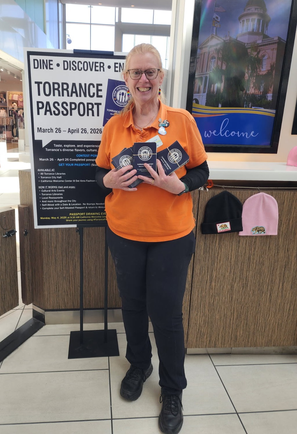 A lady in Orange shirt and black pants holding a bunch of Torrance Passport booklets at the Del Amo Mall