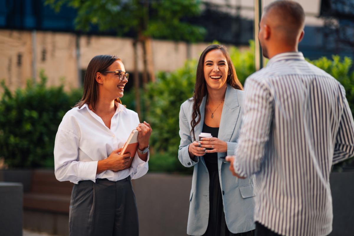 Three business people enjoying a coffee break outside their office laughing and talking in a sunny urban setting. Happy colleagues embody teamwork and friendship