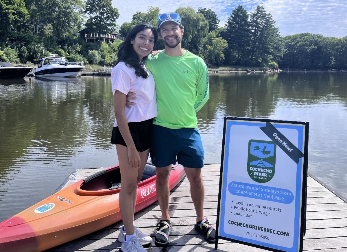 A woman and a man smile while standing on a dock next to a kayak, sign and river