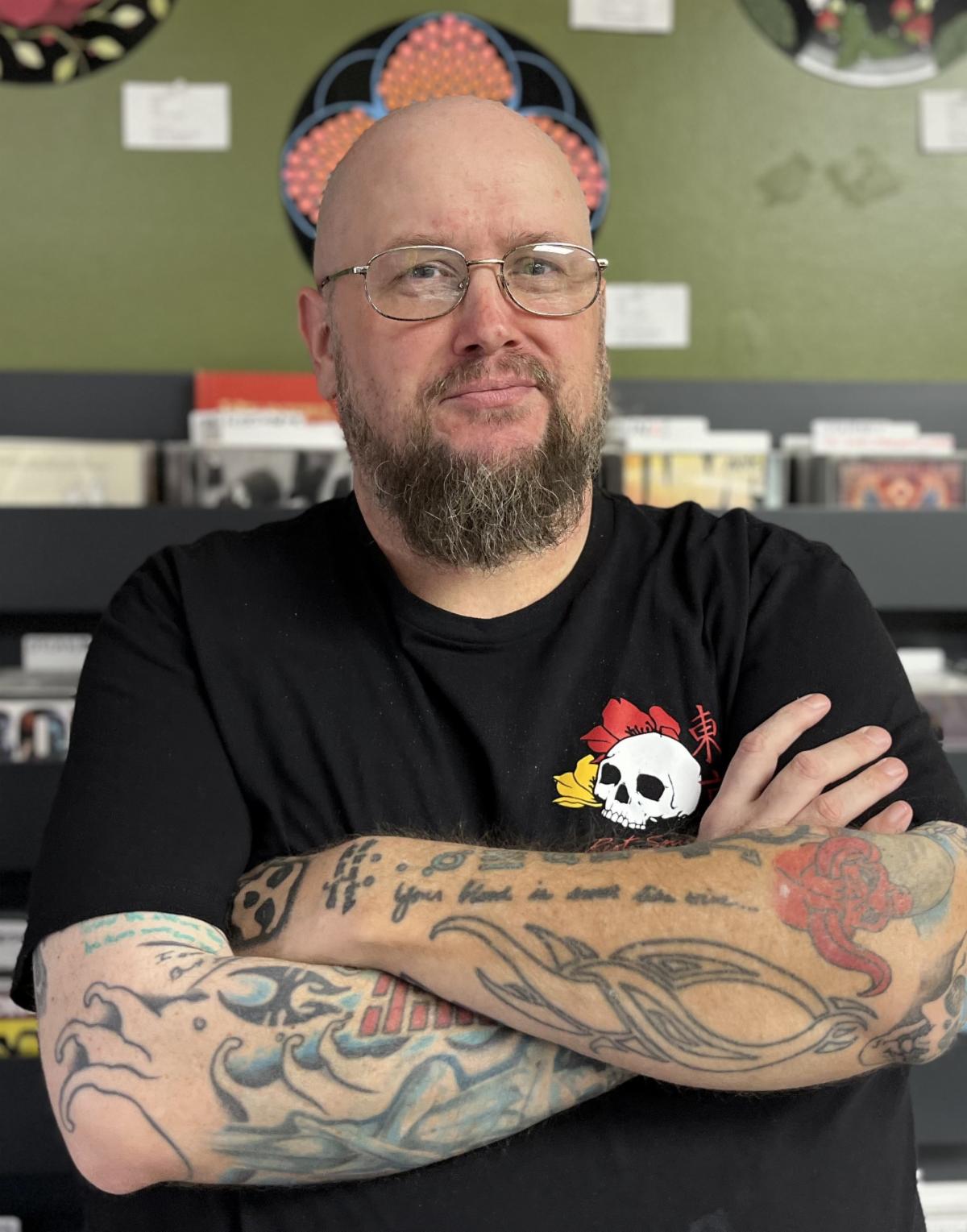 A bald man with glasses, a beard, and tattooed arms stands with folded arms in front of shelves with CDs and green wall art.