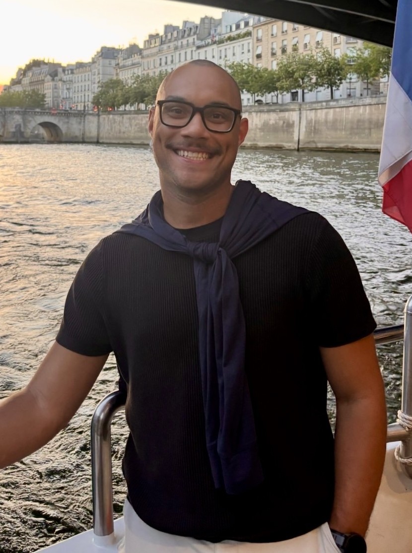 A man in a black shirt with a river in the background along with a French flag