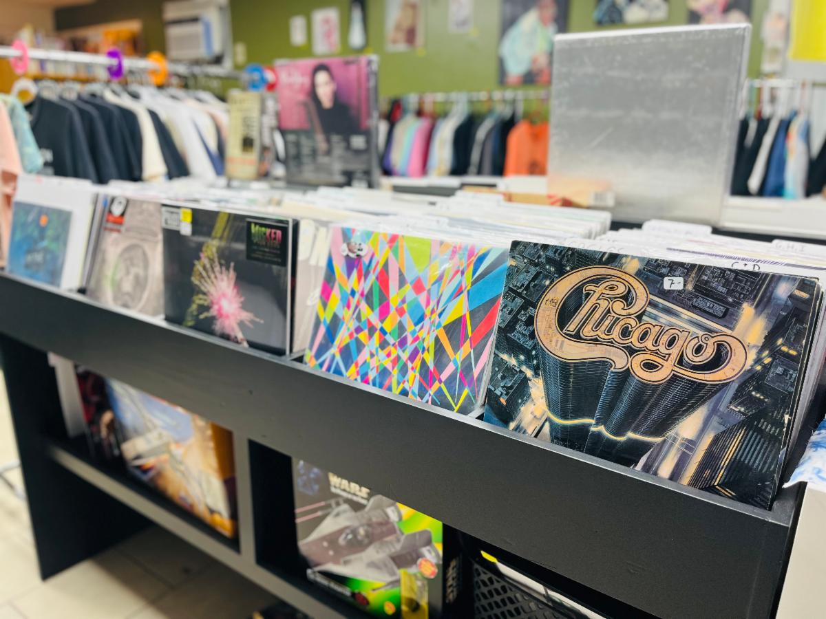 Vinyl records displayed in bins at a record store, with album covers visible, clothing racks in the background, and a colorful, well-lit interior.