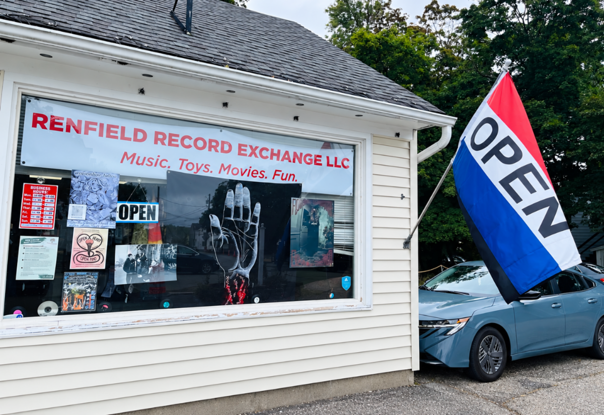 A storefront window displays â€œRenfield Record Exchange LLCâ€ and various posters. A large red, white, and blue â€œOPENâ€ flag hangs outside next to a parked car.