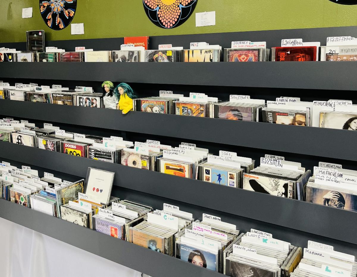 Shelves at a record store display rows of labeled CDs and album covers, with two small dolls placed among the items on the top row.