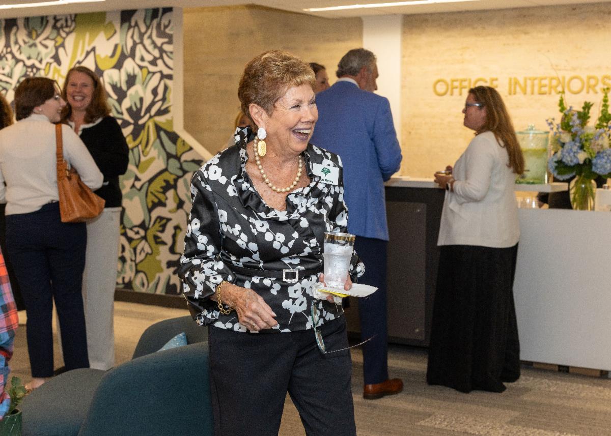 A woman in a black shirt with white flowers smiles big. In the background are people mingling.