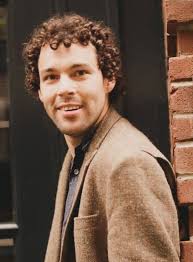 Reid with curly brown hair wearing a tweed jacket smiles at the camera.